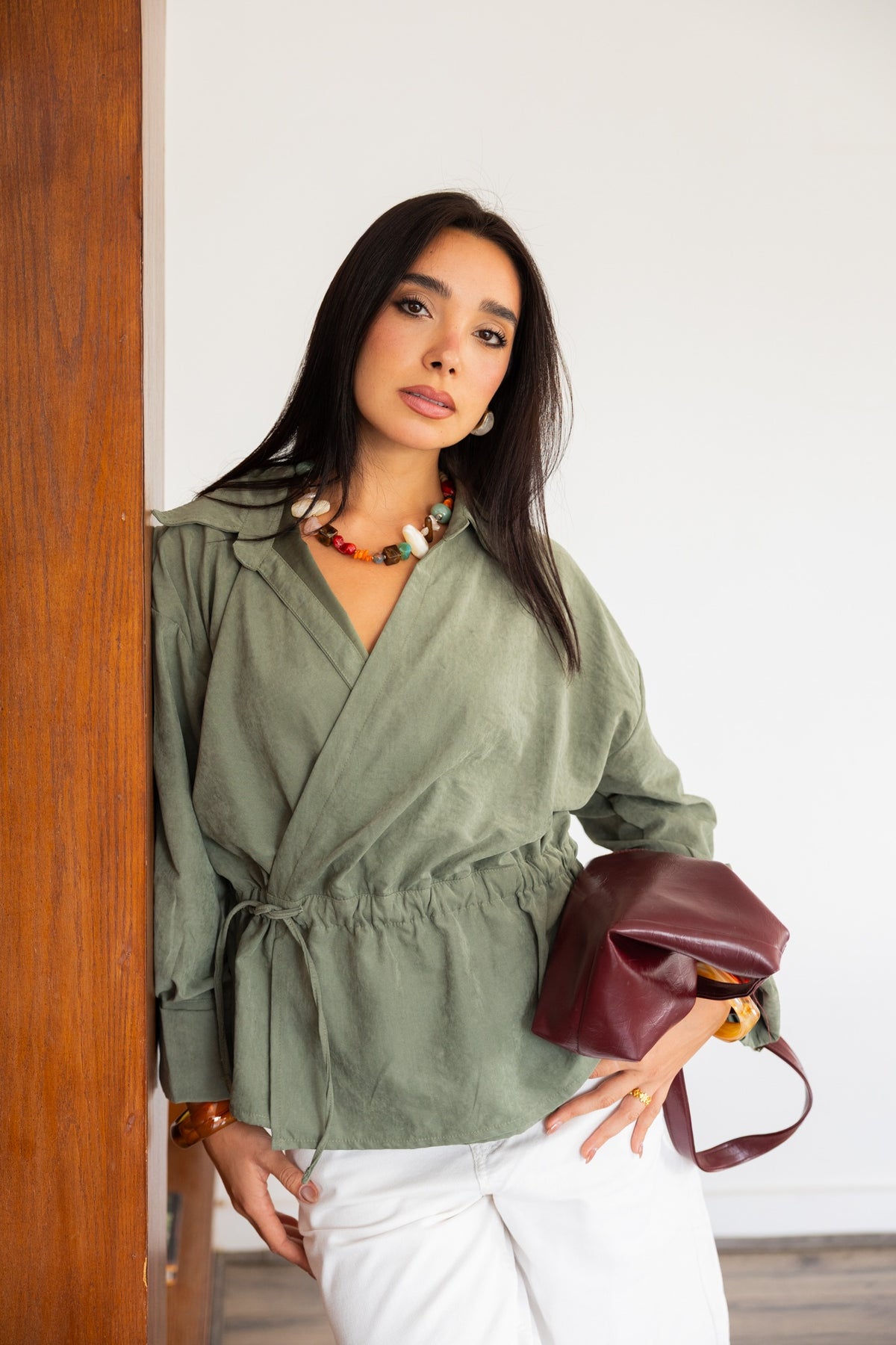 Model wearing an olive wrap blouse with white trousers and a burgundy bag